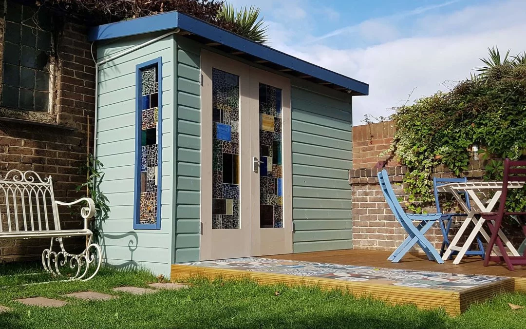 A small garden room with mosaic detail on its doors and windows sits on a wooden deck in a lush garden. Beside the shed are a white metal bench, a small round table with blue and purple chairs, and a brick wall partially covered by greenery. - a room in the garden