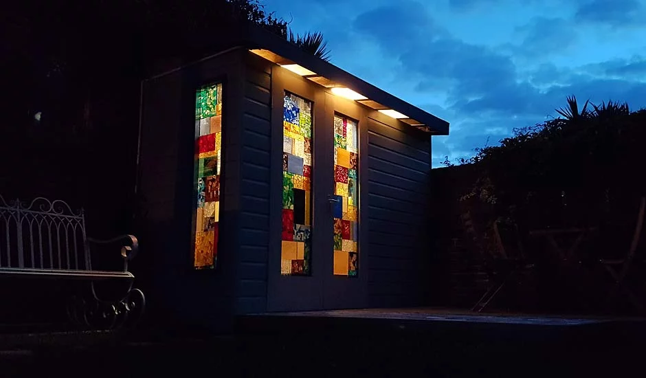 A small garden room with colorful stained glass windows is illuminated from below at dusk. The darkening sky and surrounding shadows emphasize the vibrant colors of the glass. A wrought iron bench is visible to the left, and outdoor furniture is partially hidden in the background. - a room in the garden