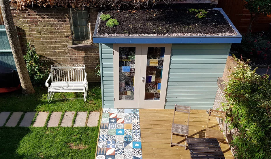 A cozy backyard scene featuring a wooden shed with a roof covered in greenery. This garden room boasts colorful stained glass windows and a door with a blue handle. In front is a tiled patio with various designs and two wooden chairs. A white bench is to the left. - a room in the garden