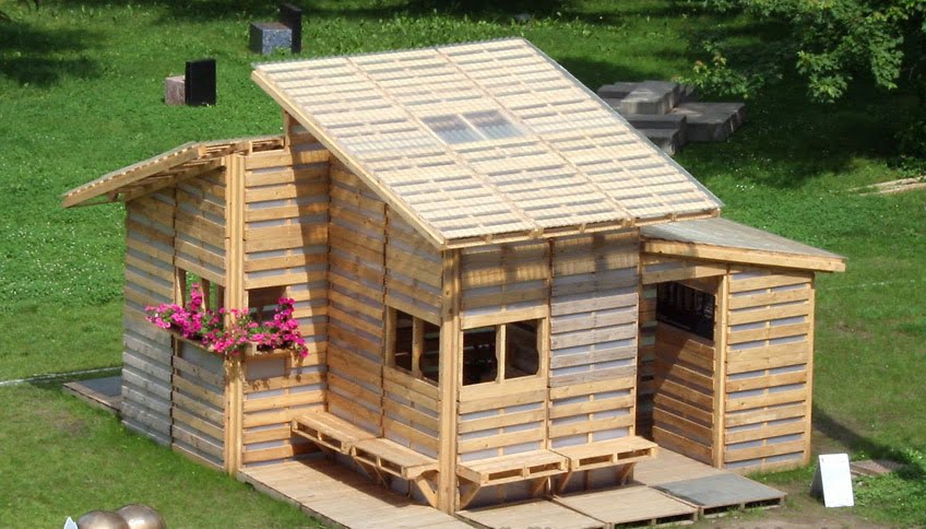 A small wooden summer house with a slanted roof and large windows sits on a green lawn. There is a flower box with pink flowers attached to the side of the house. A wooden bench is built into the front of the house, and the structure appears to be constructed from wooden slats. - a room in the garden