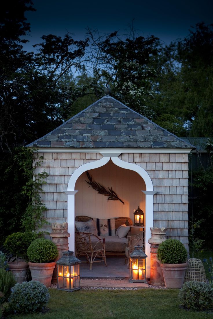 A cozy gazebo at dusk features wicker furniture with pillows and blankets. Illuminated by warm lantern light, this summer house boasts an arched entrance and is surrounded by potted plants and greenery, creating a serene and inviting outdoor space. - a room in the garden