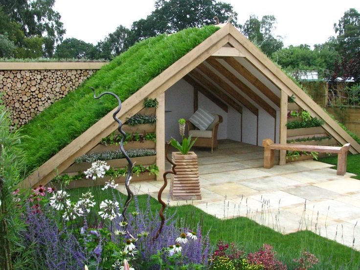 A summer house with a green roof covered in grass and plants. It has a wooden frame, open front, and sides, with a bench inside. The surrounding garden is lush with various flowers, shrubs, and a woodpile wall on the left. A winding garden sculpture is in the forefront. - a room in the garden