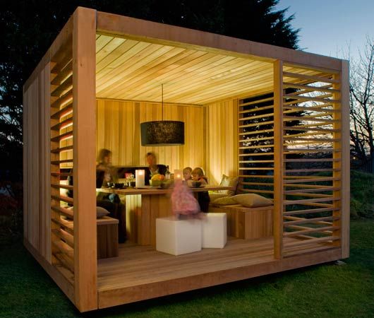 A group of people are gathered inside a modern wooden summer house. The structure features an open front, slatted sides, and ambient lighting. Inside, there’s a table with seating, and the scene appears to be during twilight in a backyard setting. - a room in the garden