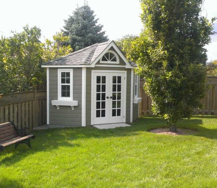 A quaint, small gray shed with white trim and French doors stands in a lush green backyard. It features a pitched roof, an arched window above the doors, and is surrounded by trees and a wooden fence. This charming summer house has a wooden bench placed on the left side near the fence. - a room in the garden