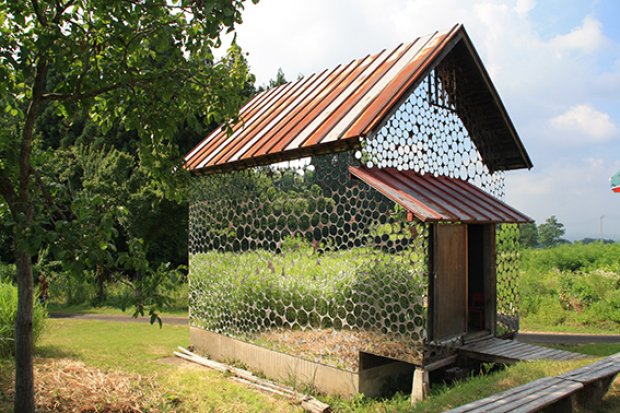 A small summer house stands in a grassy area under a clear sky; its walls are constructed from hexagonal glass bottles, creating a unique, translucent effect. The roof is made of red and white sheets of metal, and a wooden walkway leads to the entrance. - a room in the garden