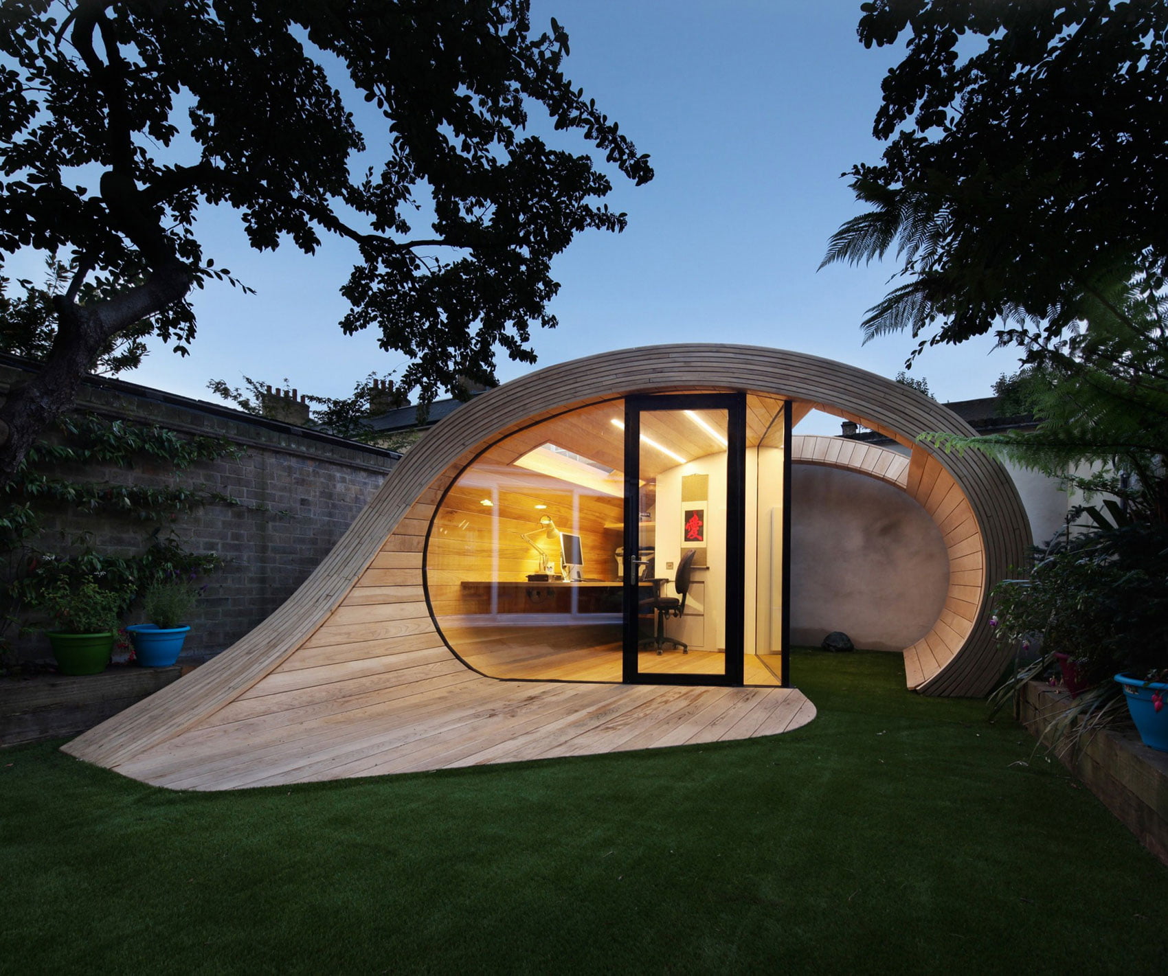 A contemporary, wooden garden office with a curved, organic design. This stylish summer house features large glass doors and windows, offering a view of the cozy interior workspace with a desk and chair. The surrounding area is beautifully landscaped with lush greenery and grass. - a room in the garden