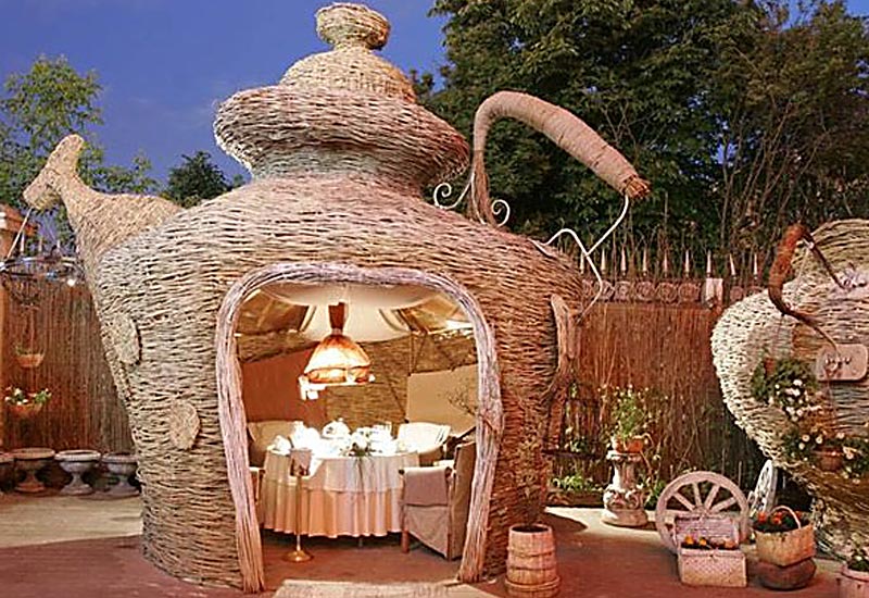 A whimsical, teapot-shaped structure made of woven materials serves as an entrance to a cozy, softly-lit summer house dining area with a circular table and chairs. Surrounding the teapot are rustic decorations, including barrels, a wheel, and a small fountain. - a room in the garden