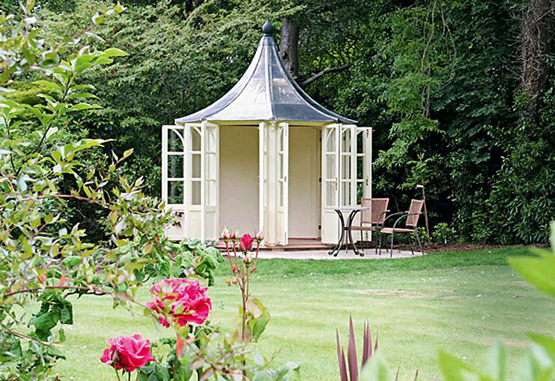 A quaint garden summer house with a black pointed roof and open double doors stands amidst lush green trees. In front, there's a small table with two chairs. The foreground features vibrant pink roses and other greenery, enhancing the serene garden setting. - a room in the garden