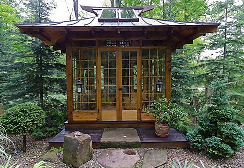 A charming summer house with large glass-paneled doors is nestled among dense greenery. It features a steep, ornate roof with decorative elements. A potted plant sits on the porch, and the surrounding area is landscaped with shrubs and stones. - a room in the garden