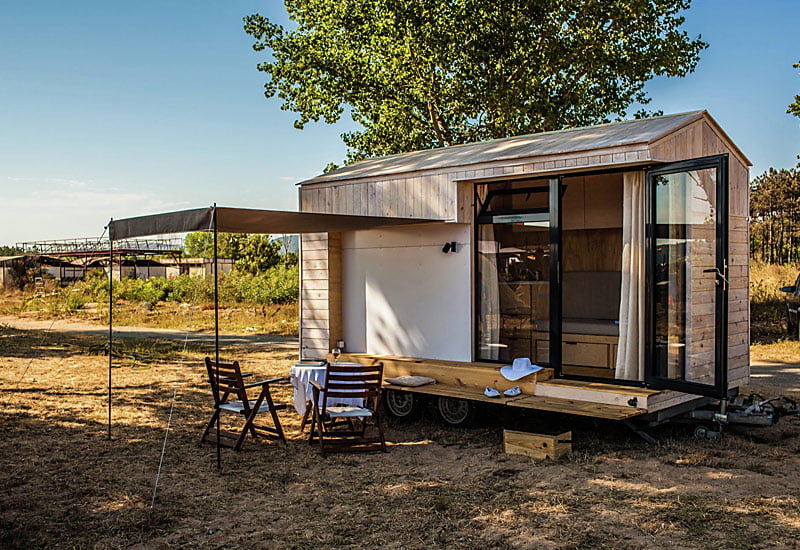 A modern tiny summer house on wheels is parked in an open, dry area with some green trees in the background. It has a glass door, a wooden porch, and an extended awning providing shade for a small outdoor dining setup with a table and four chairs. - a room in the garden