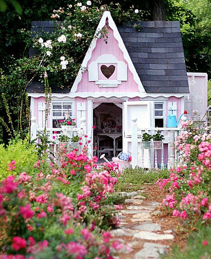 A charming, small pink playhouse with white trim and a heart-shaped window, perfect as a summer house. The house is surrounded by lush greenery and vibrant pink flowers. A stone path leads to the open front door, revealing a cozy interior with chairs just inside. - a room in the garden