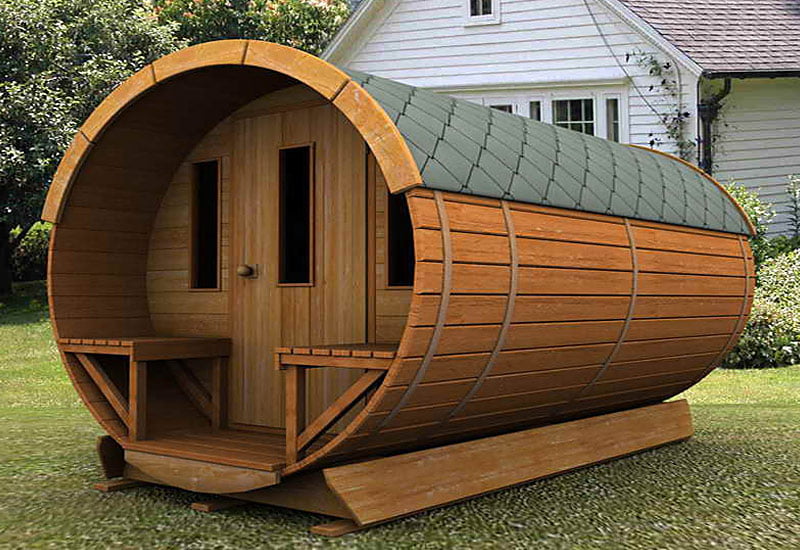 A wooden barrel-shaped mini summer house with a shingled roof, narrow windows, and a small front porch is placed on a green lawn in front of a white house with a grey roof. - a room in the garden