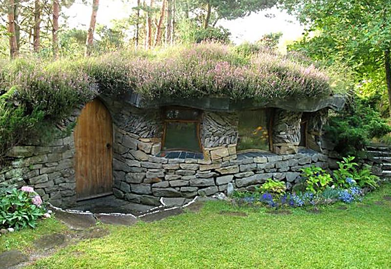 A quaint, small stone summer house with a grass and flower-covered roof nestled in a lush, green garden. The house has arched wooden doors and several irregularly shaped windows. Surrounding the summer house are various plants, trees, and a well-maintained grassy area. - a room in the garden