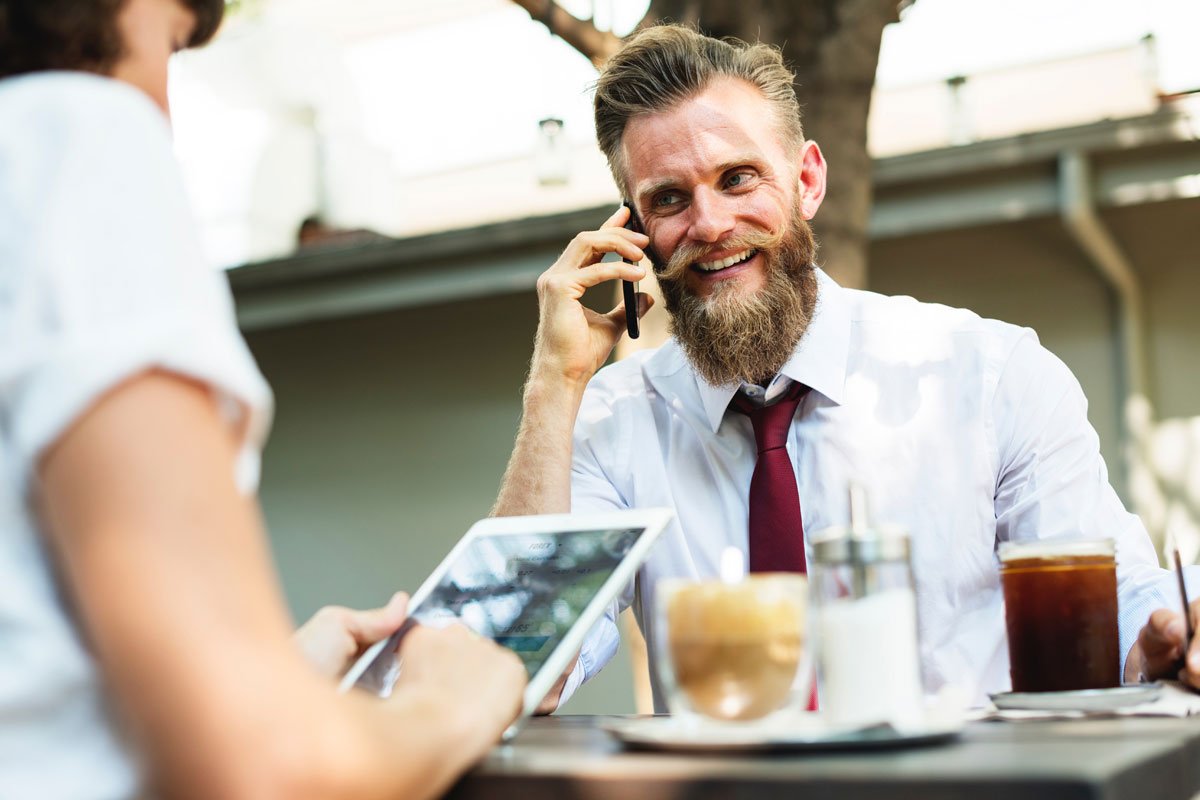 A bearded man in a white shirt and red tie is sitting at an outdoor table in a garden office, smiling while talking on a phone. In front of him are a glass of iced coffee, a latte, and a glass of water. Another person is holding a tablet and sitting across from him. - a room in the garden