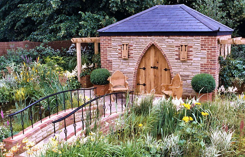 A charming garden scene featuring a small stone and brick garden office with a pointed wooden door, flanked by two chairs. It is surrounded by lush greenery and colorful flowers. A curved, wrought-iron bridge crosses a narrow pond leading to the door. - a room in the garden