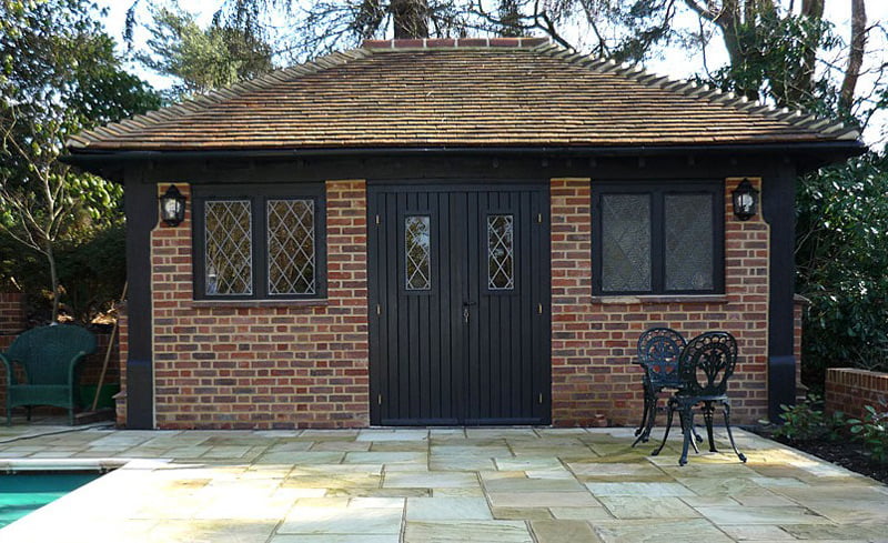 A small, brick garden shed with a tiled roof, located next to a patio with stone slabs. The shed, which might easily double as a cozy garden office, has a central wooden door flanked by windows on either side. A black wrought-iron table and two chairs are positioned on the patio in front of the shed. - a room in the garden