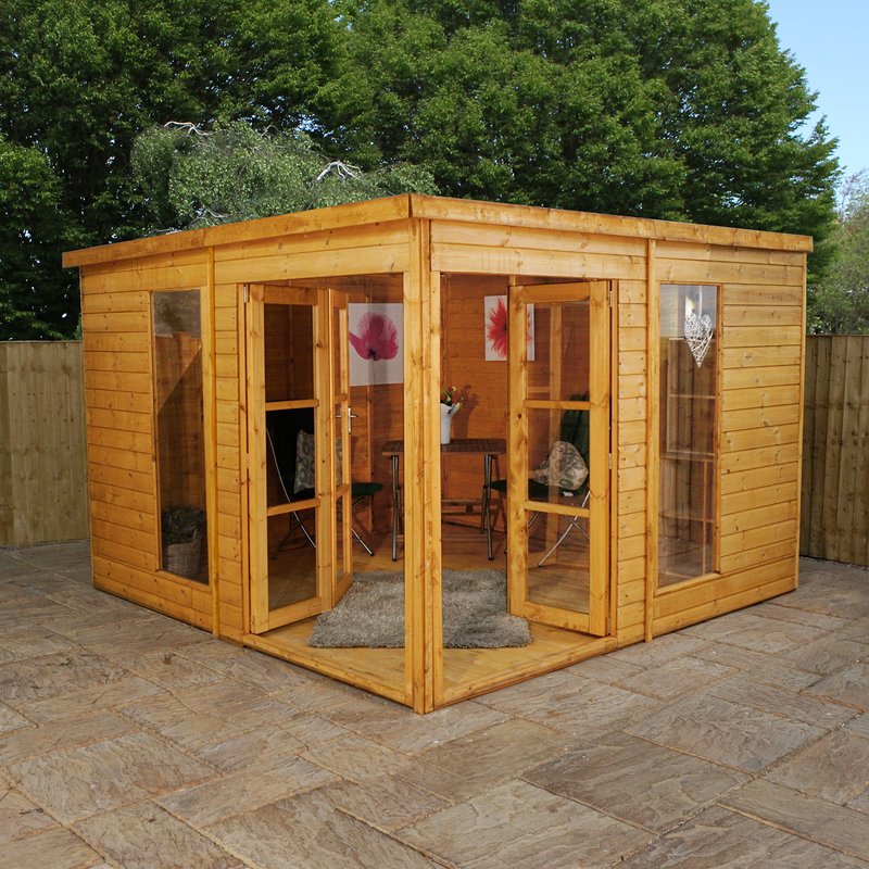 A wooden summerhouse with a double-door entrance and multiple windows stands on a paved patio area. Inside, it doubles as a garden office, featuring chairs, a small table, and framed pictures on the interior walls. The summerhouse is surrounded by a wooden fence and green trees. - a room in the garden