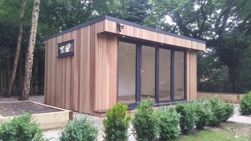 A small, modern wooden cabin with large glass doors and a narrow window on the side serves as an ideal garden office. Surrounded by greenery and trees, it features raised garden beds with plants and a paved walkway leading to the entrance. - a room in the garden