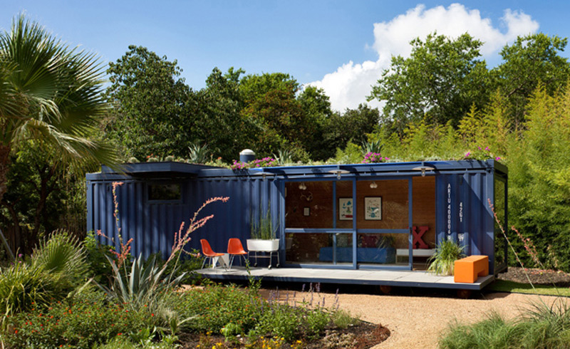 A modern blue shipping container home, ideal as a garden office, features large glass doors and windows set amidst lush greenery. The small outdoor seating area includes two orange and white chairs and a blue bench. The roof of the home has plants growing on it, enhancing the natural look. - a room in the garden