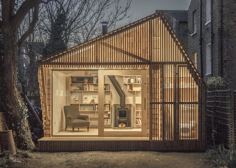 A small, modern cabin with large glass windows revealing a cozy interior. Inside, there's a comfortable armchair, bookshelves filled with books, and a wood-burning stove. The cabin doubles as an inviting garden office surrounded by trees, featuring wooden slats on the exterior. Warm evening light glows from within. - a room in the garden