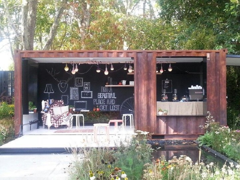 A cozy café in a converted shipping container with an open façade. The interior, reminiscent of a garden office, features chalkboard art on dark walls, hanging lights, a small sofa, and a few stools. Outside, there's lush greenery and wildflowers, creating a serene, inviting atmosphere. - a room in the garden