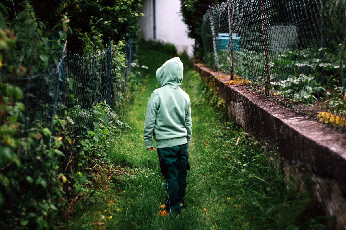 A child wearing a green hoodie and dark pants stands on a narrow path between two fences, surrounded by lush greenery. The child faces away, looking towards a house in the distance, possibly pondering how planning permission shaped this secluded and tranquil landscape. - a room in the garden