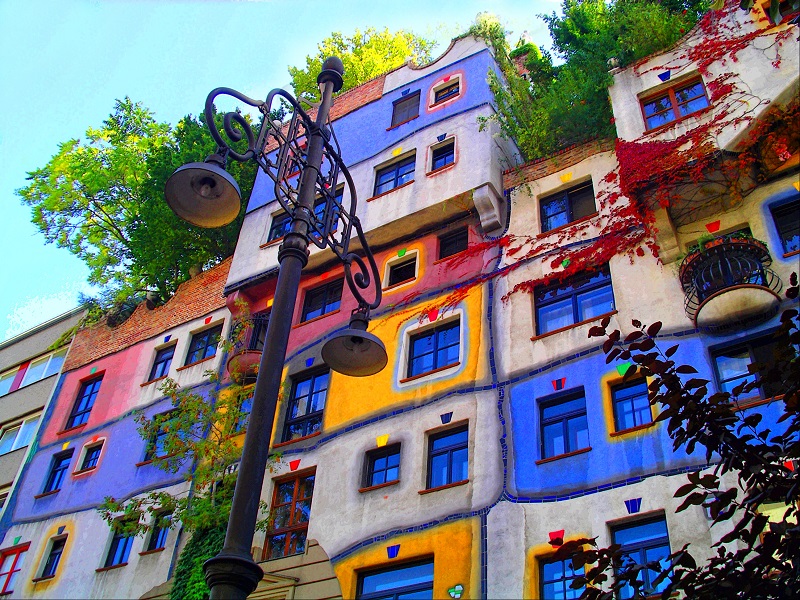 A multicolored building with a whimsical exterior features a variety of vibrant hues such as blue, yellow, and red. The architecture is adorned with plants and greenery, creating an artistic and lively atmosphere. A vintage streetlamp stands prominently in the foreground. - a room in the garden