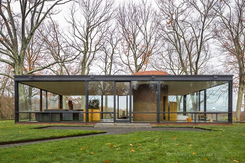 A modern glass house with large transparent walls stands in an autumnal landscape surrounded by leafless trees. The architecture highlights minimalistic furnishings and an industrial chimney, with a pathway leading up to the front of the building. - a room in the garden