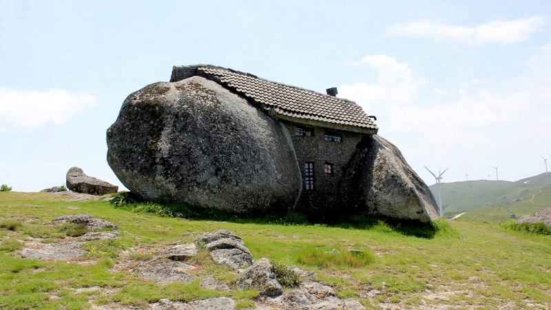 A unique stone house built between large boulders is situated on a grassy hill. The structure's architecture features a tiled roof and small windows, blending seamlessly with the natural rock formations. In the distant background, wind turbines are visible under a partly cloudy sky. - a room in the garden