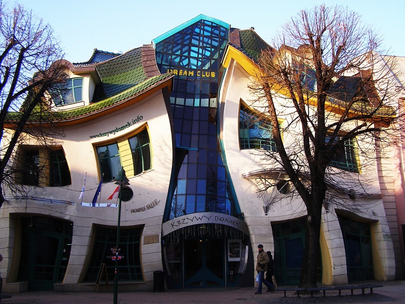 A whimsical, wavy-structured piece of architecture with a distorted façade, known as the Krzywy Domek (Crooked House), located in Sopot, Poland. The building features curved walls and windows, a blue glass entrance, and green-shingled roofs. Trees frame the front. - a room in the garden