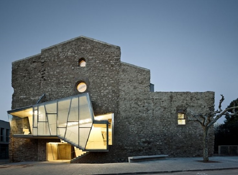 A stunning piece of architecture, the modern structure with its angular, glass entrance extends from the grey stone facade of an old building. The juxtaposition of the contemporary design against the ancient structure is highlighted in the evening light. A leafless tree stands to the right. - a room in the garden