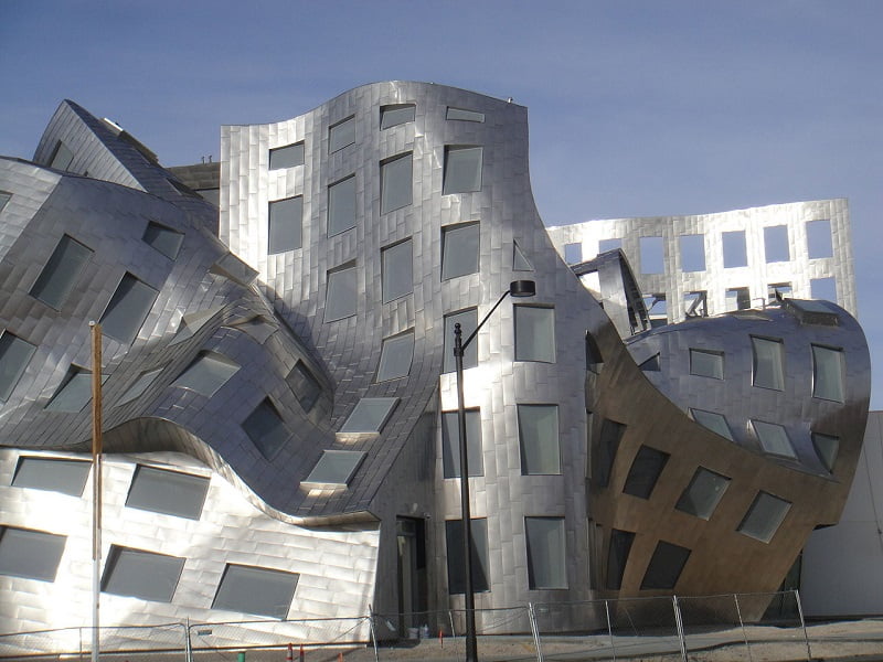 A modern building featuring a distinctive, warped exterior made of shiny, metallic panels and irregularly shaped windows. The structure's undulating design gives it a dynamic, wave-like appearance. A pole and fence are visible in front of this stunning piece of architecture. - a room in the garden