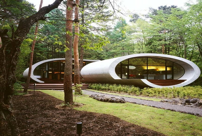 A marvel of modern architecture, this elliptical-shaped house with large glass windows is nestled in a lush forest. The structure features curved, tunnel-like forms with wood steps leading to the entrance. The surrounding area is abundant with tall trees and greenery, creating a serene escape. - a room in the garden