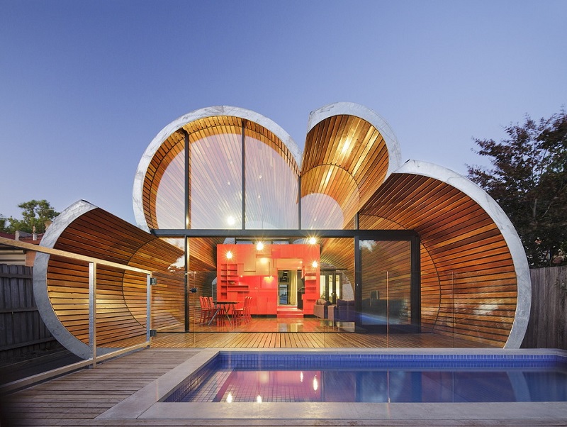 A modern house with a unique clamshell roof design featuring curved wooden panels and large glass windows showcases stunning architecture. Inside, vibrant red furniture and a sleek kitchen are visible. The exterior includes a swimming pool in the foreground with decking surrounding it. - a room in the garden