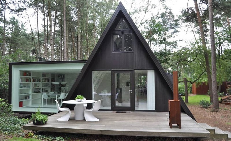 A marvel of modern architecture, this A-frame house with a large glass extension is nestled in a wooded area. The extension features a wall of bookshelves and a white table with chairs. A wooden deck in front of the house contains a small round table with white chairs and potted plants. - a room in the garden