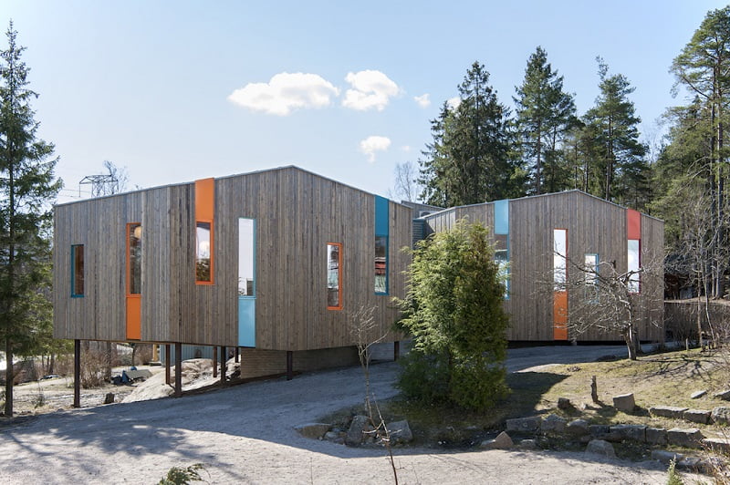 A modern two-story building with a wooden exterior stands elevated on stilts over a gravelly terrain, surrounded by tall trees. The architecture features narrow, vertical windows with colorful accents in orange, blue, and yellow. The sky is partly cloudy. - a room in the garden