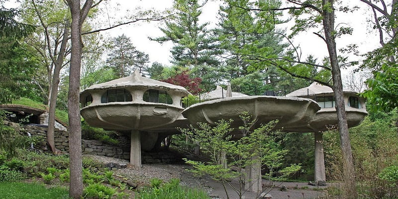 An unusual piece of architecture, this house consists of two circular, UFO-like structures elevated on pillars nestled among trees. The building has a futuristic design with large windows and is surrounded by wooded greenery. A path leads up to the entrance of one structure. - a room in the garden