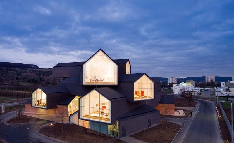 A modern architectural building at dusk featuring stacked gabled structures with illuminated, large windows. The surrounding landscape includes a road and green spaces, with a view of distant hills and a cloudy sky in the background, showcasing the epitome of contemporary architecture. - a room in the garden