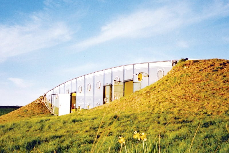 A modern piece of architecture, this partially underground building features a curved glass façade and circular windows, set against a backdrop of clear blue sky. Integrated into a grassy hillside, it blends naturally with the landscape. Yellow flowers adorn the foreground, enhancing its beauty. - a room in the garden