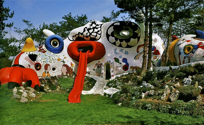 A large, colorful sculpture resembling a fantastical creature with various patterns and shapes, including a red slide that forms its tongue. The architecture of the sculpture is surrounded by greenery, trees, and rocks, enhancing its whimsical and imaginative appearance. - a room in the garden