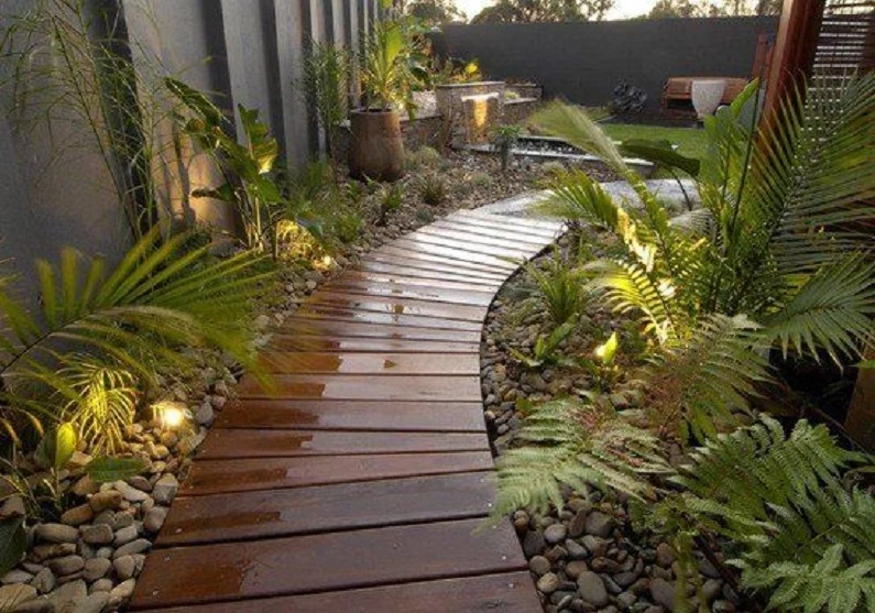 A narrow wooden pathway winds through a garden adorned with green plants, ferns, and illuminated by ground lights. River rocks line both sides of the path, creating a serene, natural ambiance. This charming setup offers inspiring path ideas with a cozy seating area visible in the background. - a room in the garden