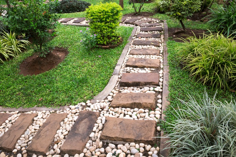 A garden pathway made of rectangular stone slabs with white pebbles in between offers excellent path ideas. The pathway winds through green grass and various plants and shrubs. The stones are arranged in a linear fashion, and the garden surroundings are neat and well-maintained. - a room in the garden