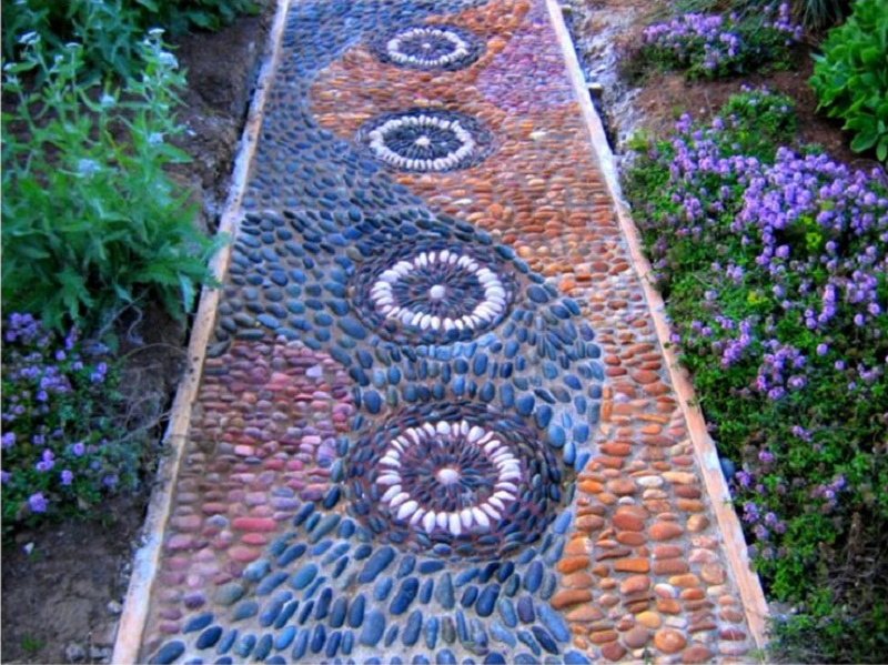 A garden pathway, perfect for path ideas, is decorated with a mosaic of small stones in an intricate pattern. The mosaic features concentric circles of black and white stones, surrounded by swirls of blue and orange stones. Lush green plants and small purple flowers border the path. - a room in the garden