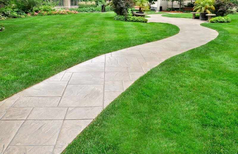 A winding paved pathway curves through a well-manicured lawn in a garden, offering charming path ideas. The path is surrounded by lush green grass, various plants, and flowers lining the edges. In the background, there are trees, shrubs, and a couple of benches near the greenery. - a room in the garden