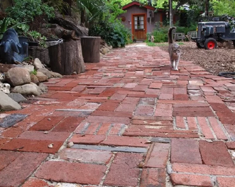 A cat walks down a brick path towards a small house with a red door, surrounded by greenery and garden items. Logs and baskets are seen on the left side of the path, while a rustic chair and an old tractor part adorn the right, offering charming path ideas for any countryside setting. - a room in the garden