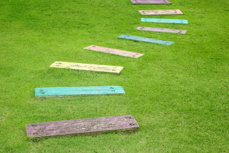 Colorful wooden stepping stones arranged in a zigzag pattern across a grassy lawn. The planks are painted in various shades, including blue, yellow, and purple, contrasting with the vibrant green grass—perfect for creative path ideas. - a room in the garden