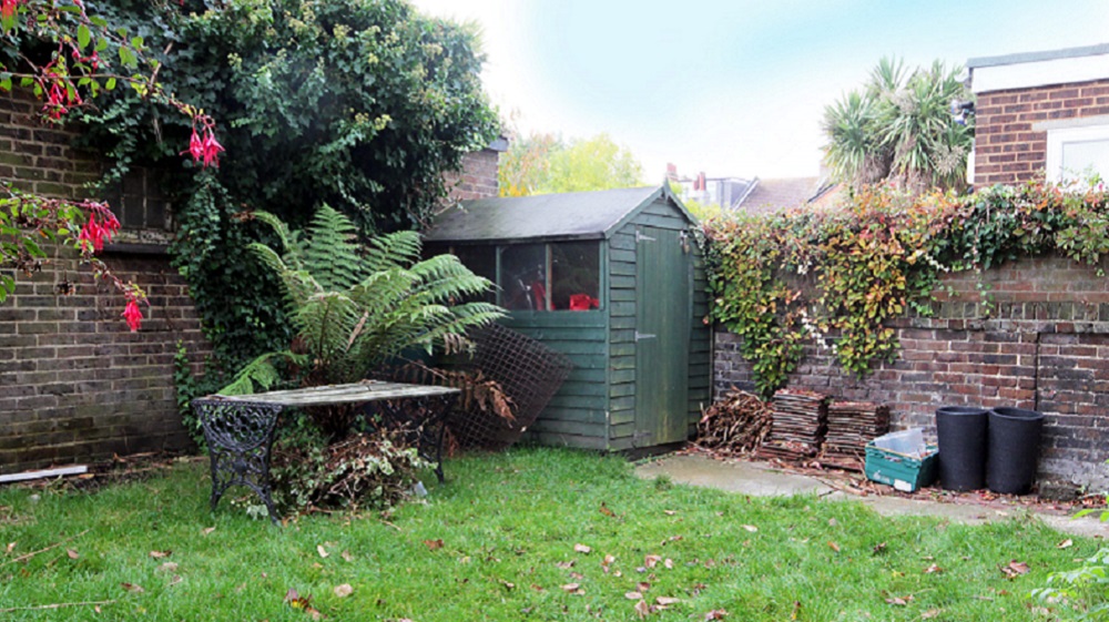 A garden scene features a small green storage room, a wooden table with a metal chair, and various plants including a fern. The space is enclosed by brick walls covered with ivy. Stacks of firewood and a few black planting pots sit near the shed. - a room in the garden