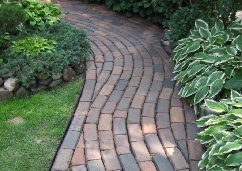 A winding brick pathway, one of many enchanting path ideas, is bordered by lush greenery on both sides. Neatly trimmed bushes with variegated leaves are seen on the right side, while smaller green plants and grass occupy the left. The pathway curves gently, creating a peaceful garden scene. - a room in the garden