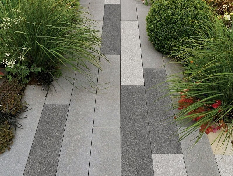 A pathway made of rectangular grey and black tiles is flanked by various green plants and shrubs on both sides. The alternating tile shades create a modern, geometric pattern. These path ideas highlight how lush greenery adds a natural contrast to the sleek, contemporary design of the path. - a room in the garden