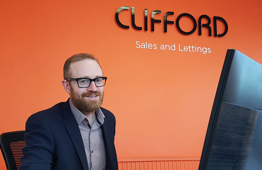 A bearded person wearing glasses and a suit sits at a desk in front of an orange wall with the company name "CLIFFORD" and the words "Sales and Lettings" displayed, reminiscent of an organized storage room. A part of a computer monitor is visible in the foreground. - a room in the garden